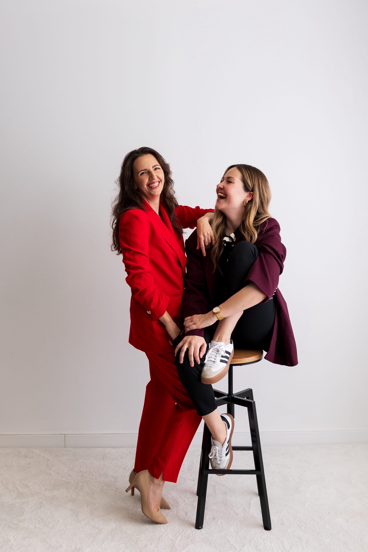 Demi en Mandy lachend samen, Demi in een rood pak, Mandy op een kruk